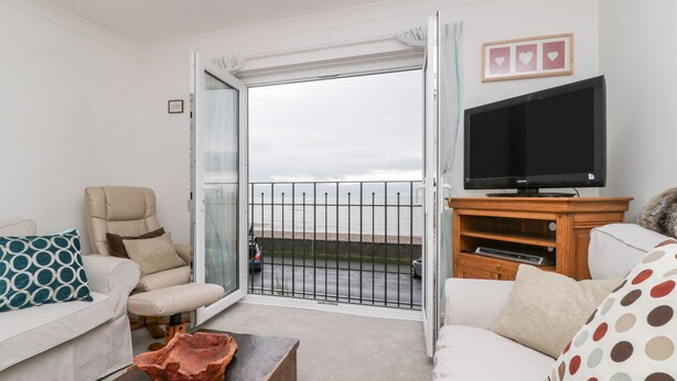 A living room with a sofa, armchair, and a coffee table. There's a TV on a wooden stand and a large window with a view of the sea. - 14 Lyme Mews (Seaton)