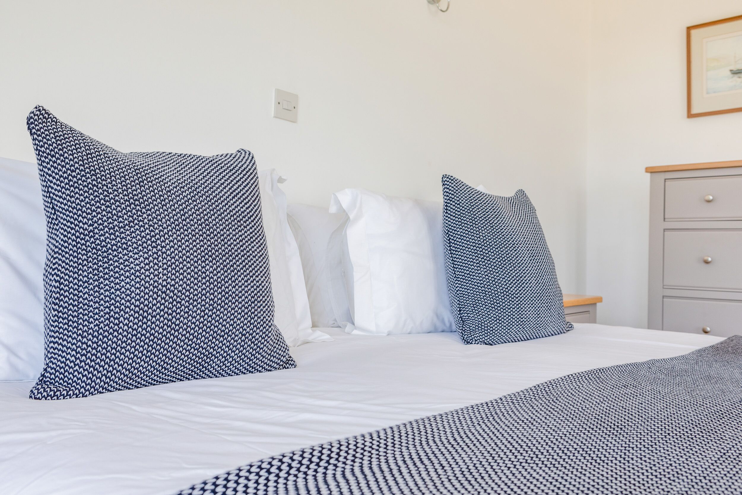 A neatly made bed with white and patterned pillows, a grey dresser, and a framed picture on the wall.