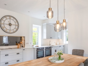 A modern kitchen with white cabinetry, a large wall clock, pendant lights, and a wooden dining table with a vase of flowers.