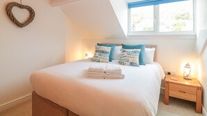 Un dormitorio con cama, almohadas, una mesita de noche, una lámpara, un adorno en forma de corazón en la pared y una ventana con cortinas azules.