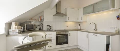 A modern kitchen with white cabinetry, a built-in oven, a microwave, and a sink. There is a chair with a high backrest and a countertop with a kettle and books.
