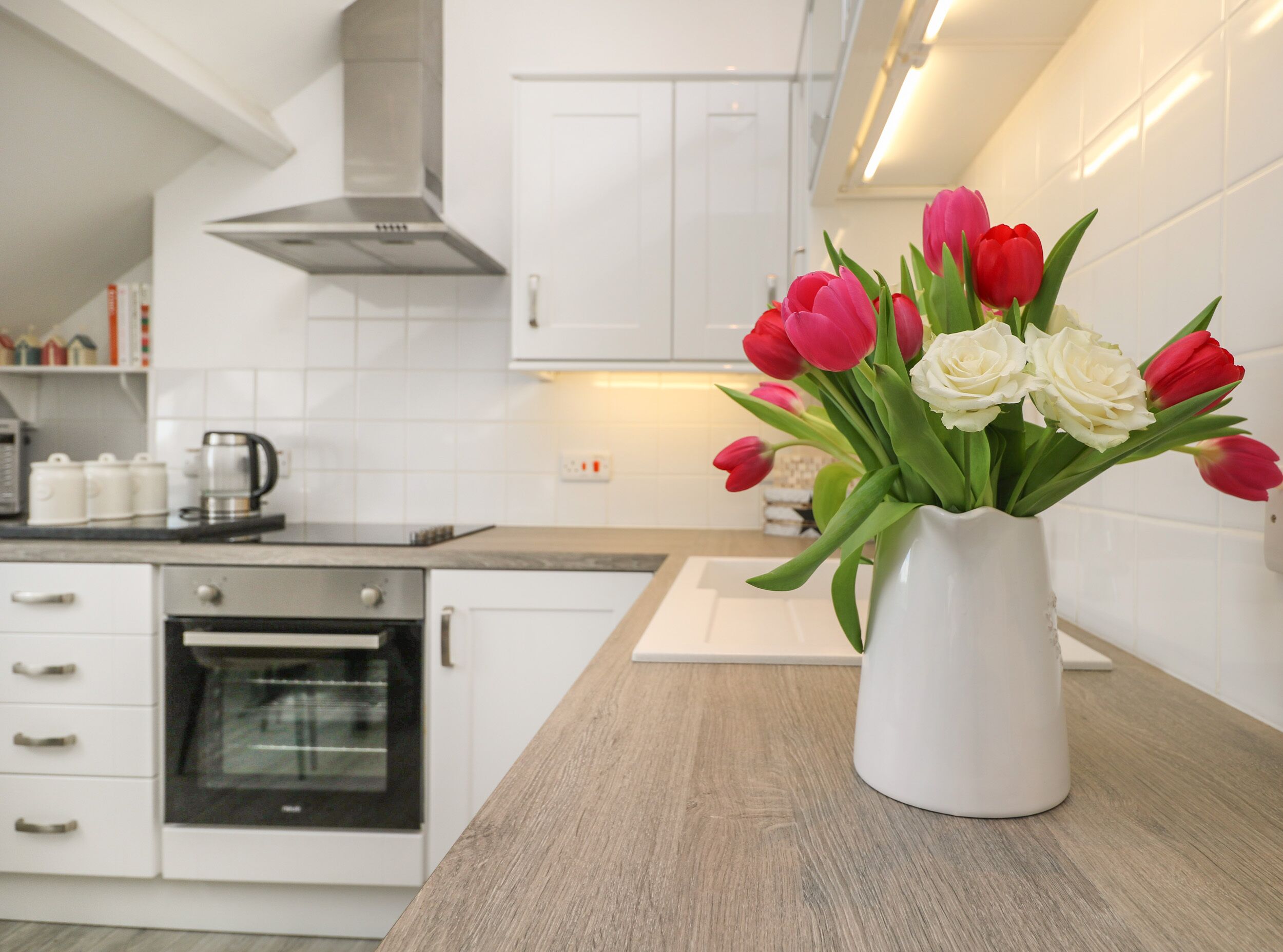 Una cucina moderna con mobili bianchi, una cappa aspirante in acciaio inossidabile e un vaso bianco con tulipani rossi e bianchi sul piano di lavoro.
