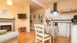 A cozy living room with a fireplace, a dining area with a table set for two, and a kitchen with white cabinets and a black range hood. - The Roost (Camborne)