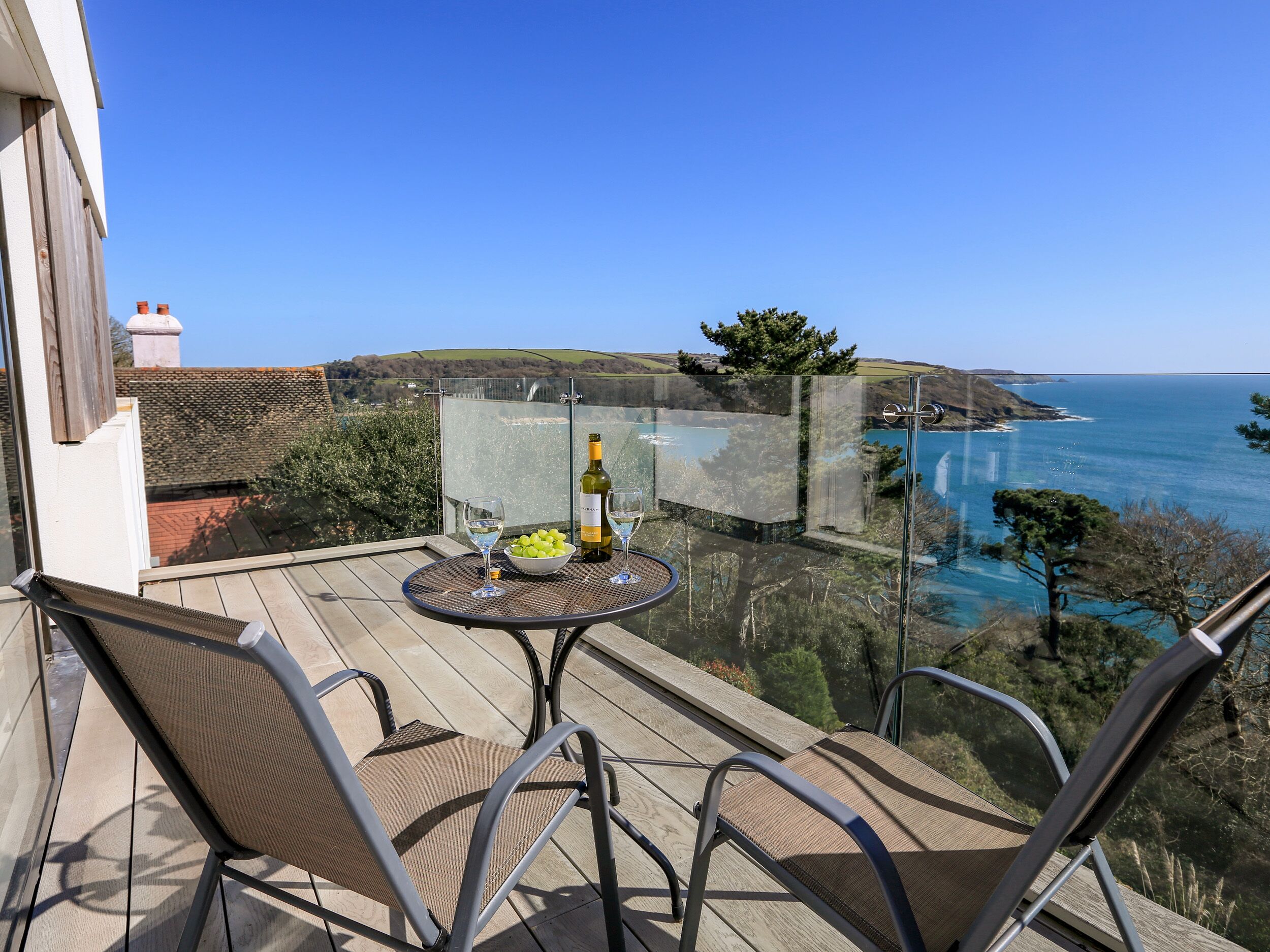 A balcony with a table, chairs, and a bottle of wine, overlooking a coastal landscape.