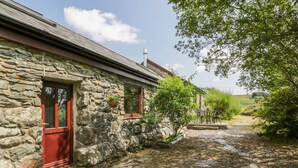 A stone building with a red door and windows, a small garden with a table and chairs, and a stone pathway leading to the entrance. - Melin Bach (Garndolbenmaen)