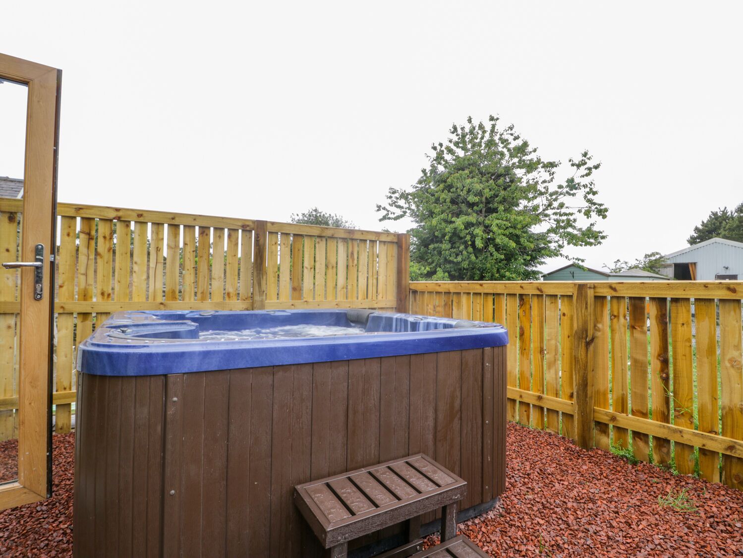 A wooden hot tub with a bench in front, surrounded by a wooden fence and mulch.