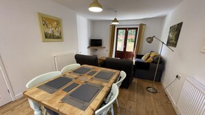 A dining area with a wooden table and chairs, a sofa, a television, and a radiator. - Stable Cottage (Winscombe)