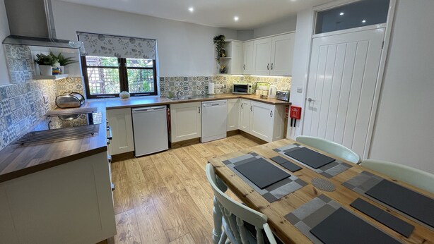 A modern kitchen with white cabinets, a wooden countertop, and a dining table with placemats. - Stable Cottage (Winscombe)
