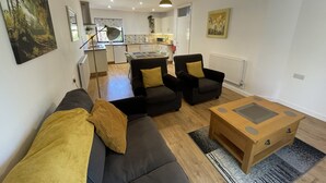 A modern living room with a sofa, armchairs, and a coffee table. There is a kitchen in the background with a dining table and chairs. - Stable Cottage (Winscombe)