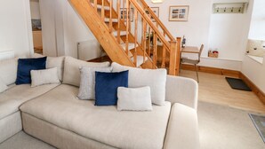 Una sala moderna con una escalera de madera, un sofá con cojines, una zona de comedor y una cocina al fondo.