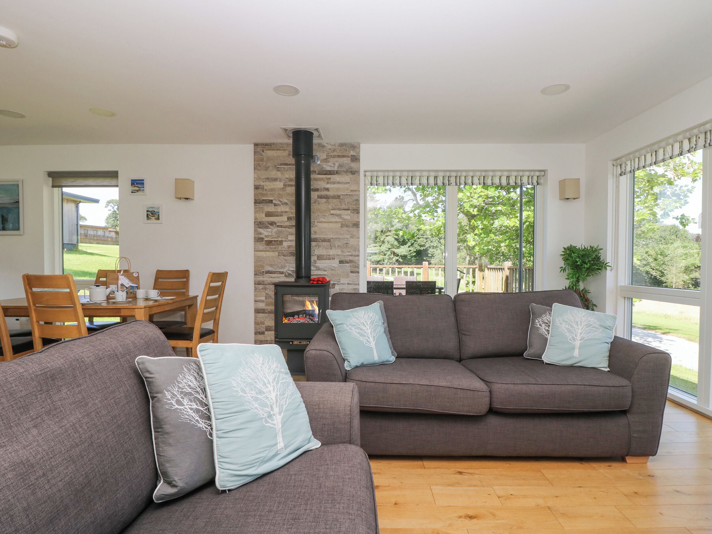 A cozy living room with a stone fireplace, a dining area with wooden furniture, and large windows offering a view of the outdoors.