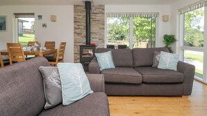 A cozy living room with a stone fireplace, a dining area with wooden furniture, and large windows offering a view of the outdoors. - Willow (Truro)