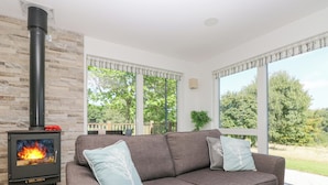 A cozy living room with a fireplace, a grey sofa, and large windows offering a view of the outdoors. - Willow (Truro)