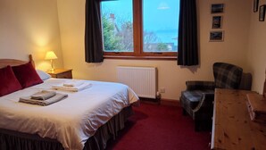 Ett hotellrum med en säng, en stol, ett bord, ett fönster med gardiner och ett element.