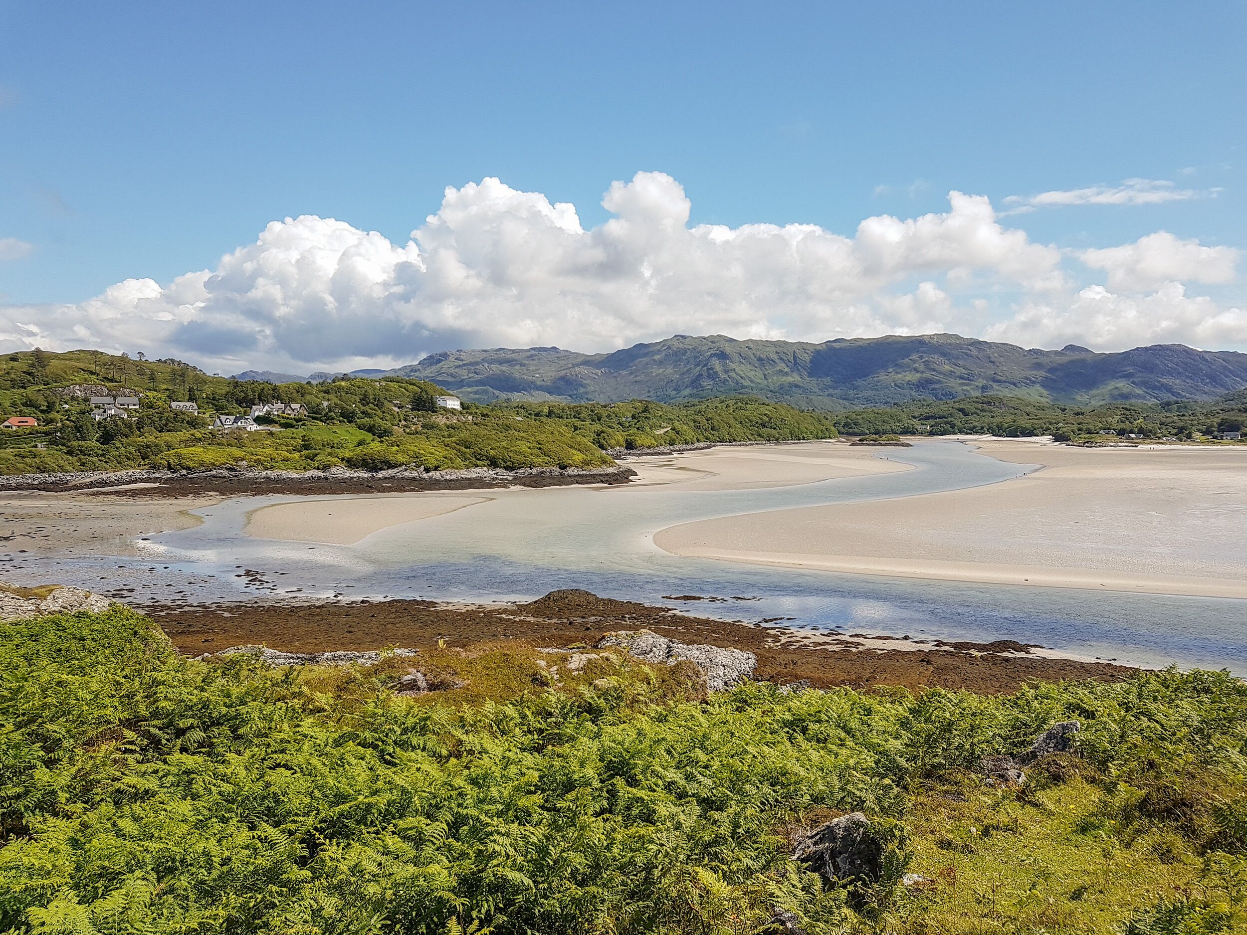 Et kystlandskab med en sandstrand, en rolig bugt og grønne bakker og bjerge i baggrunden.