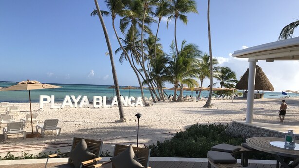 Un patio frente a la playa con sillones y una mesa, con vista a una playa de arena, palmeras y el océano.