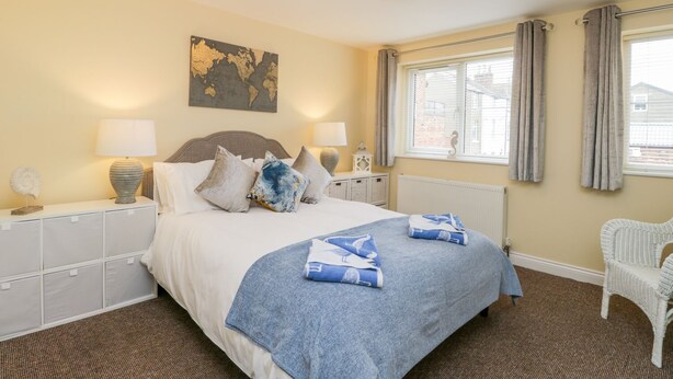 A neatly arranged bedroom with a large bed, two bedside tables with lamps, a world map artwork, a window with curtains, and a wicker chair. - Valiant House (Scarborough)