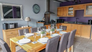 A dining area with a wooden table set for six, surrounded by grey upholstered chairs. A clock is mounted on the wall above the table. - Royal Oak (Matlock)
