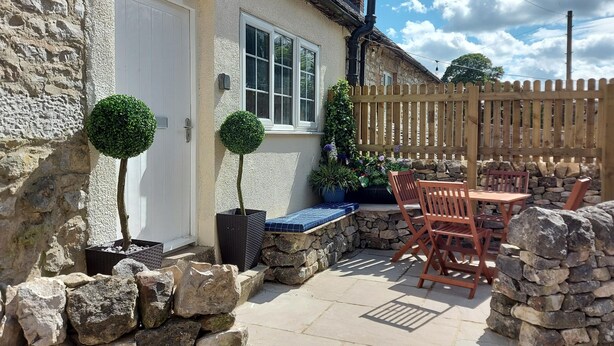 A stone house with a white door, a window, and a wooden fence. There is a patio with a table and chairs, and potted plants. - Royal Oak (Matlock)