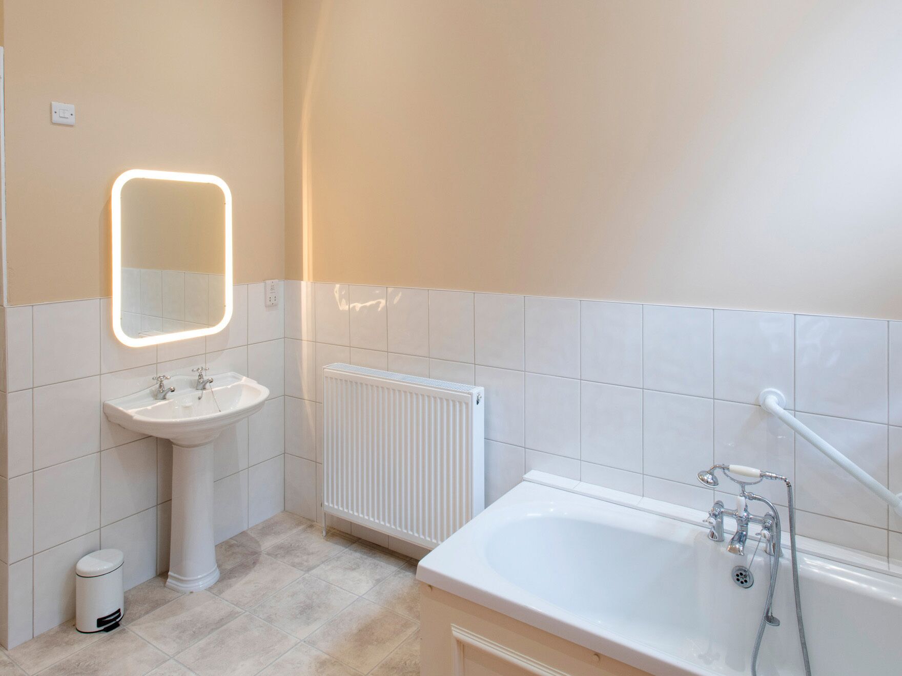 Un bany modern amb un lavabo de pedestal, un mirall amb il·luminació integrada, un radiador blanc i una banyera amb plat de dutxa.