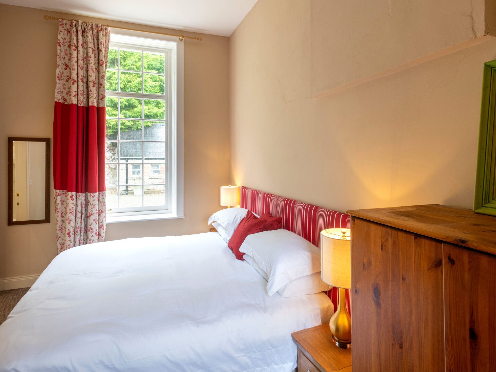 Una cama bien hecha con sábanas blancas, una cortina roja con estampado floral y una mesita de noche de madera con una lámpara.