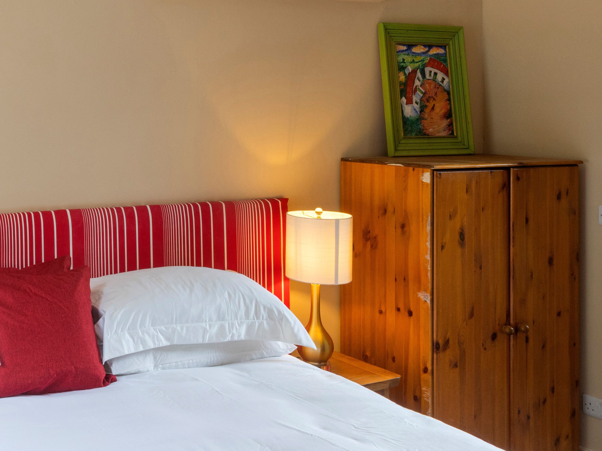 Habitación de hotel con cama, almohadas, mesita de noche con lámpara, armario de madera y un cuadro en la pared.