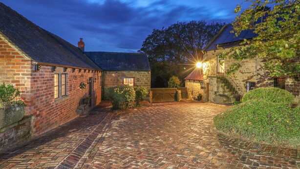 A cobblestone courtyard with stone buildings, a brick building with a window, and a tree. - Woodland Lodge (Alfreton)
