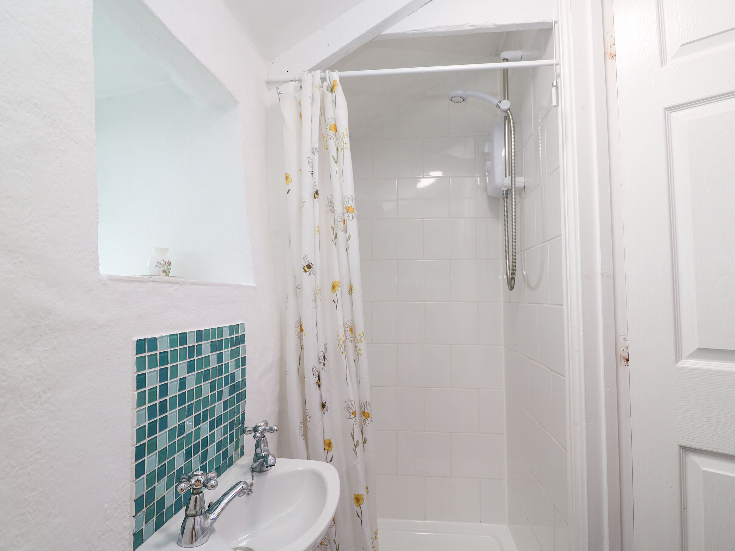 Un baño con un lavamanos blanco, una ducha con cortina de motivos florales y una ventana pequeña con un florero.