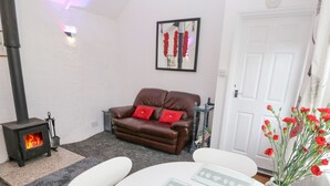 A cozy living room with a fireplace, a brown leather sofa with red cushions, a white table with a fruit bowl, and a vase with red flowers. - Toadstools (Helston)