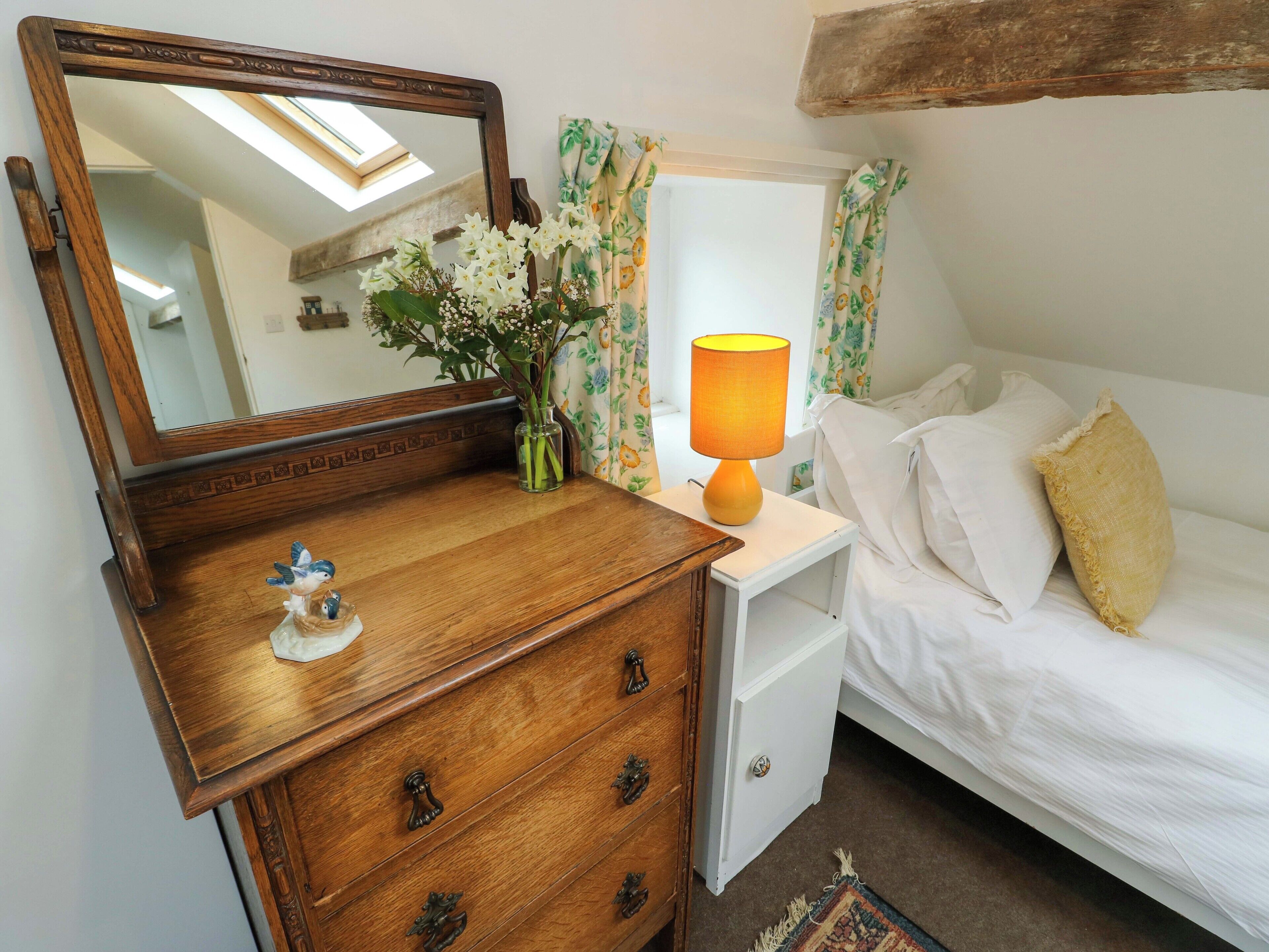 A bedroom with a wooden dresser, a nightstand with a lamp, a bed with white bedding, and a mirror reflecting the room.