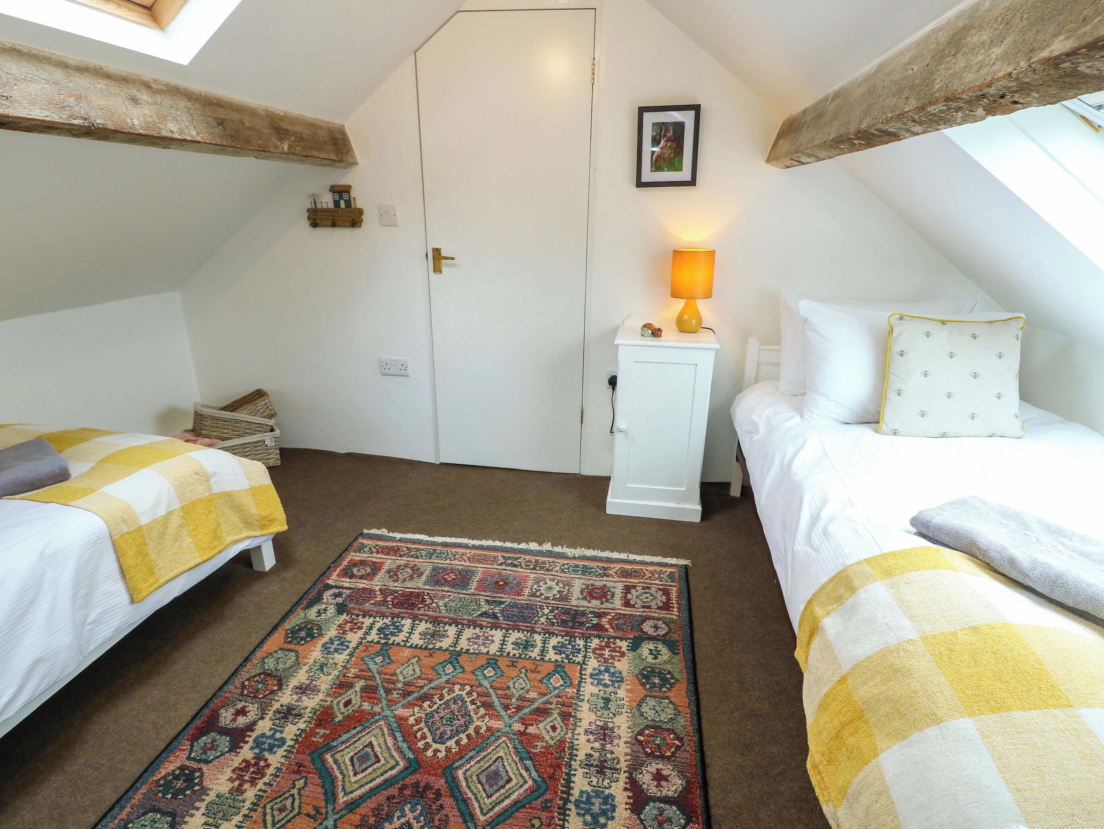 A cozy attic bedroom with two beds, a patterned rug, a small white cabinet with a lamp, and a framed picture on the wall.