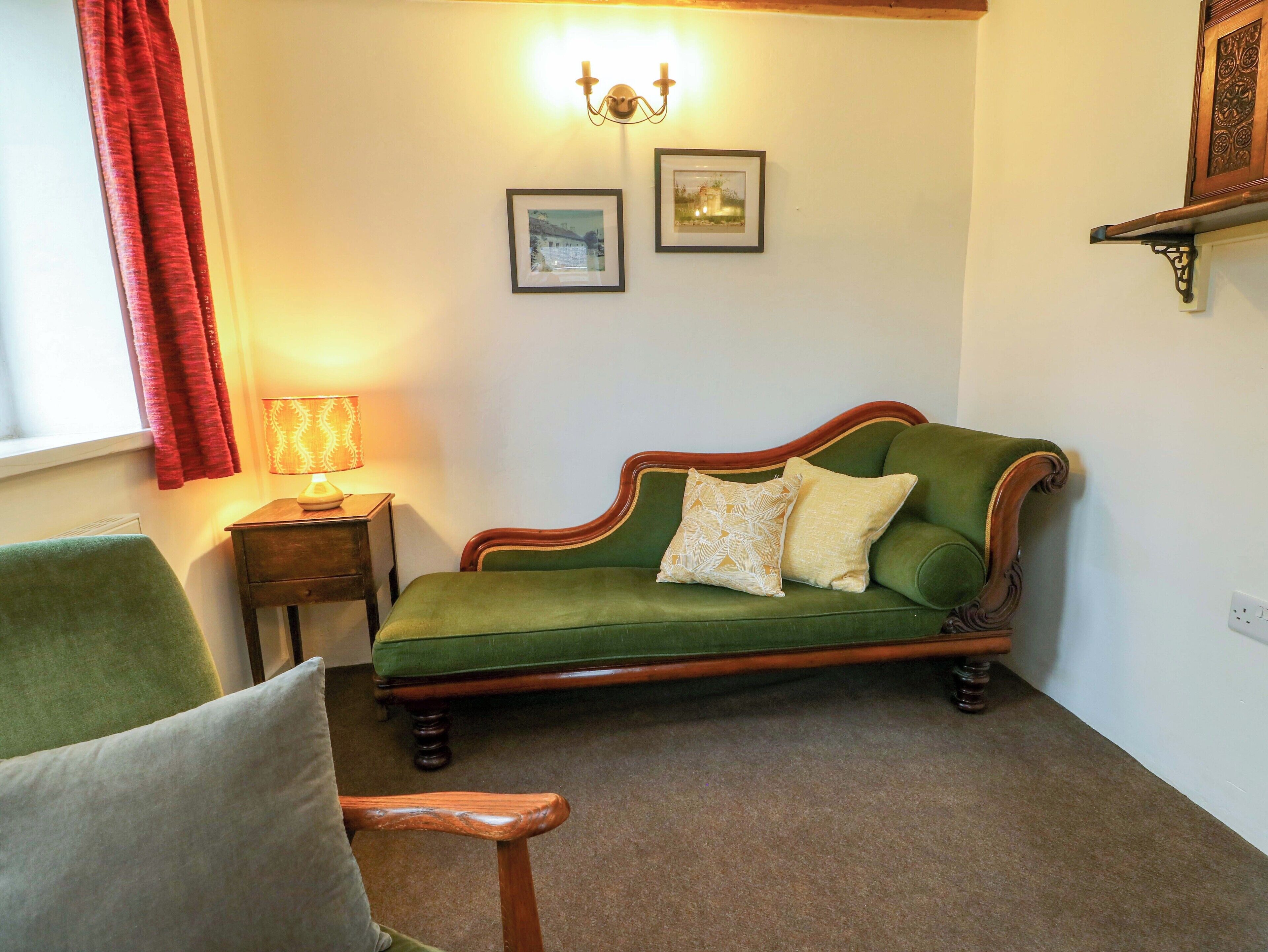 Un canapé vert avec deux coussins, une petite table d’appoint en bois avec une lampe, et deux tableaux encadrés accrochés au mur.