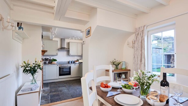 A well-lit kitchen with white cabinets, a dining table set for two with plates, glasses, and a bottle of wine. A window with curtains is visible, and a small plant is on the table. - Rigg Cottage (Whitby)