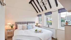 A bedroom with a large bed, a chandelier, a window with a view, and a bedside table with a lamp. - Rigg Cottage (Whitby)