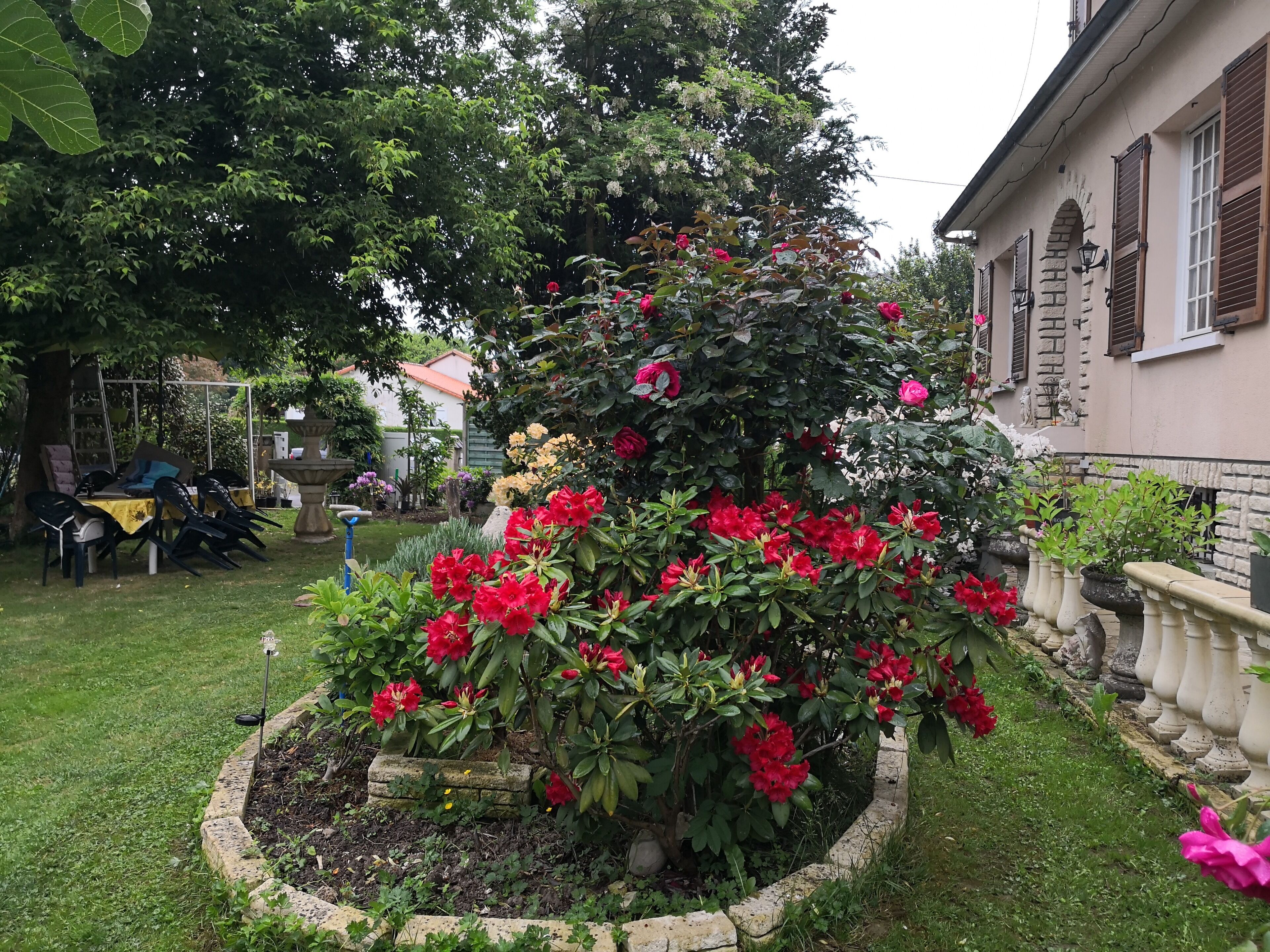 Un jardín con diversas plantas y flores, una fuente de piedra y mobiliario para exterior.