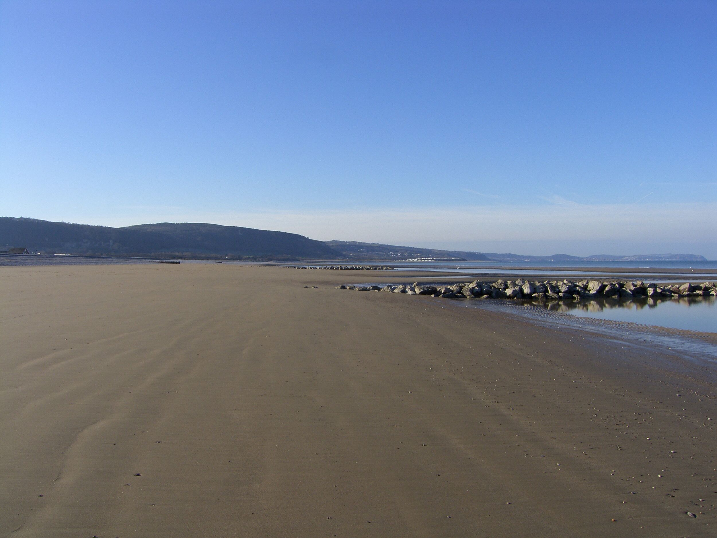 Uma praia extensa e arenosa, com um paredão rochoso e colinas ao longe.