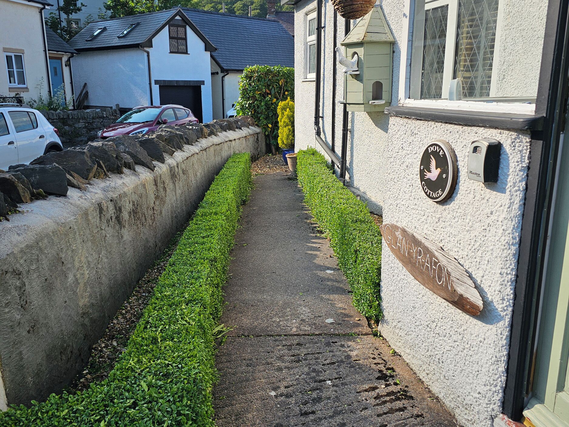 A narrow pathway flanked by a stone wall and trimmed hedges, leading to a building with a sign reading "GLAN-YR-AFON".
