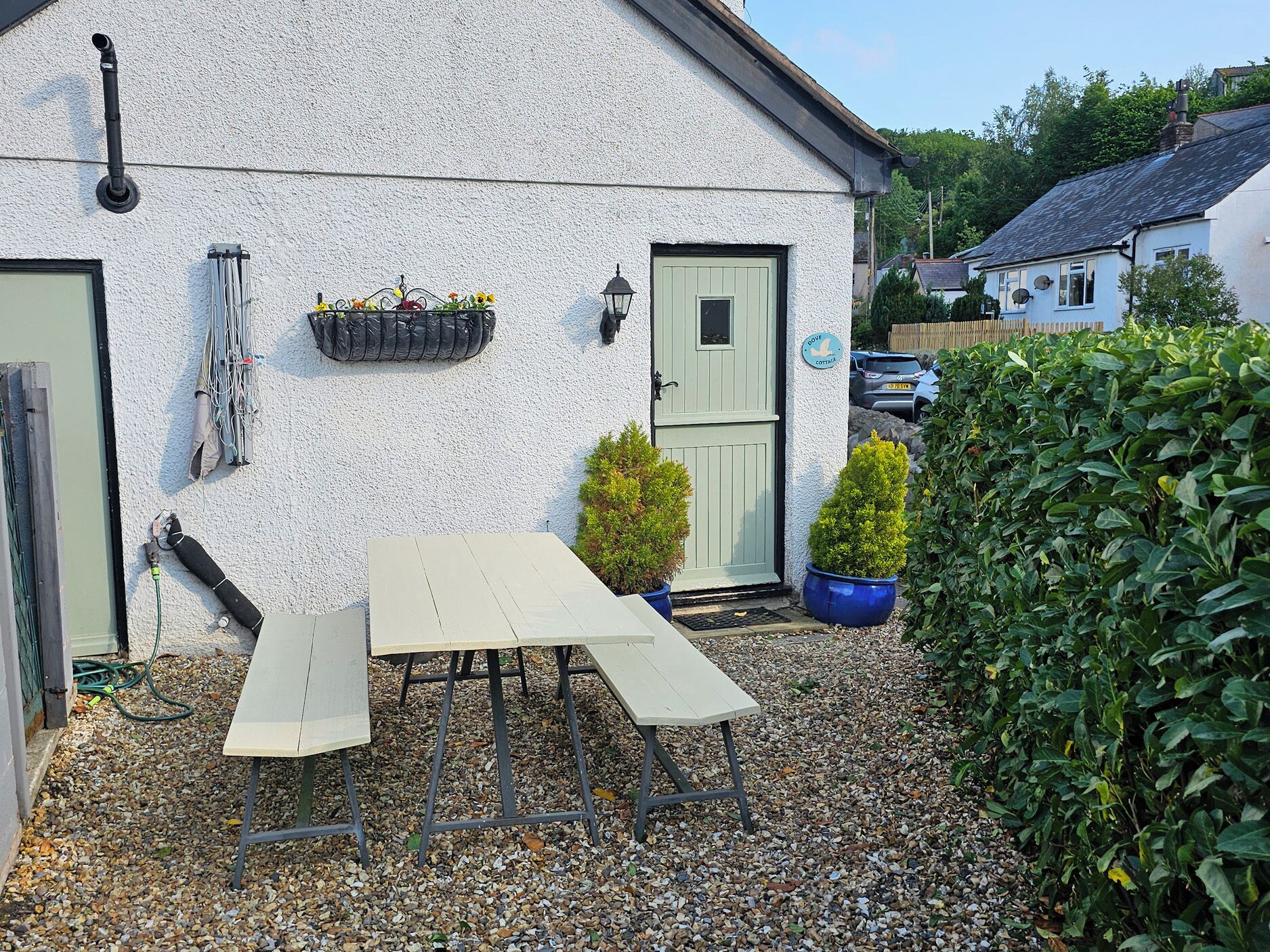 Une petite maison blanche avec une porte verte, une petite table de jardin et des plantes en pot.