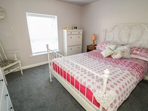 Ein Schlafzimmer mit weißem Metallbettgestell, einer rot-weißen Bettdecke, einem weißen Schminktisch, einem Nachttisch mit Lampe, einem Sessel und einem Fenster mit Jalousien.