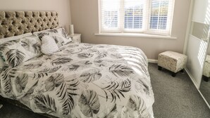 A bedroom with a tufted headboard, a bed with a floral-patterned bedspread, a nightstand with a lamp, and a window with blinds. - Cranny Corner (Ballymena)