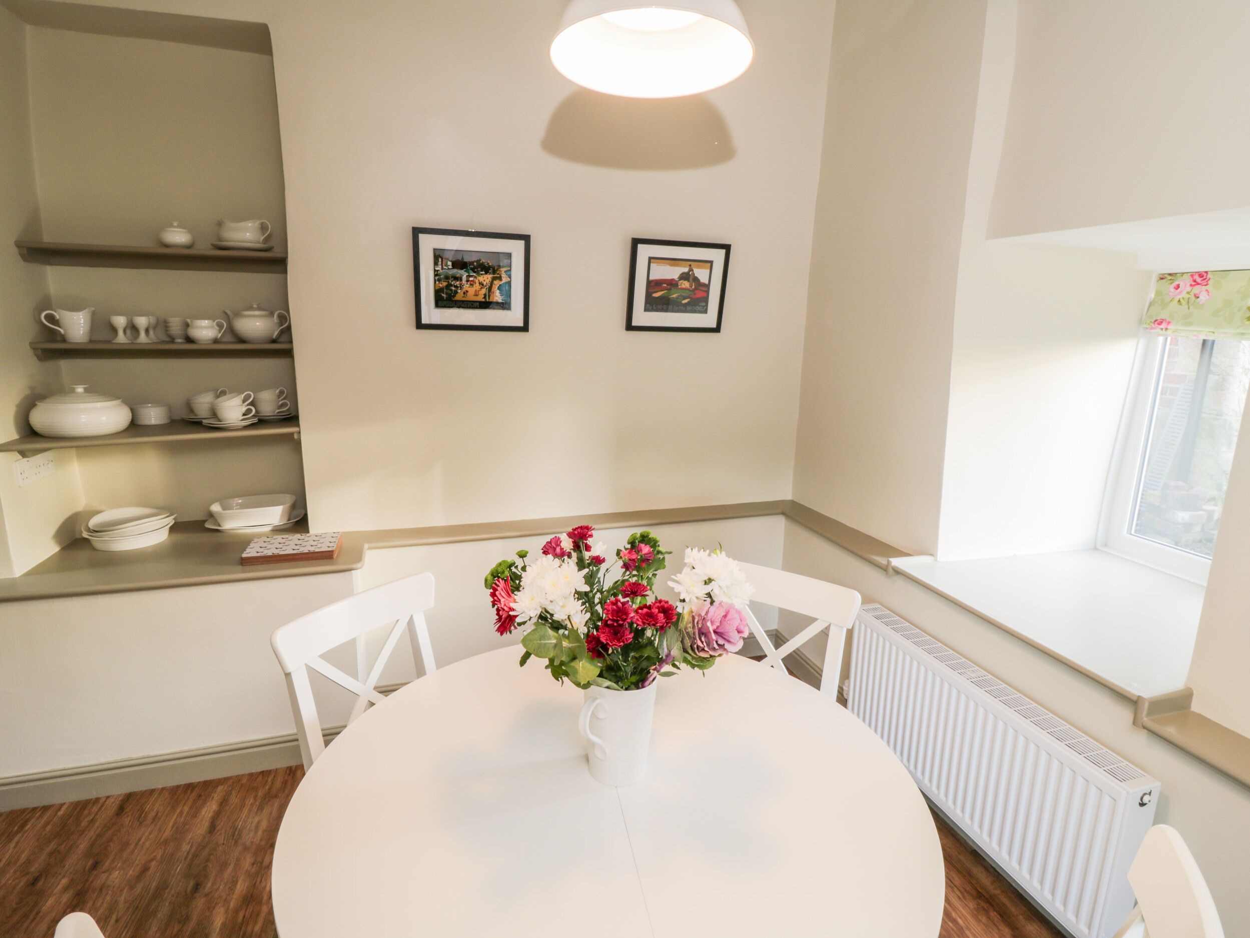 Comedor con mesa redonda, sillas blancas, un jarrón con flores y estanterías con vajilla blanca. Hay dos cuadros en la pared y una ventana con vistas.