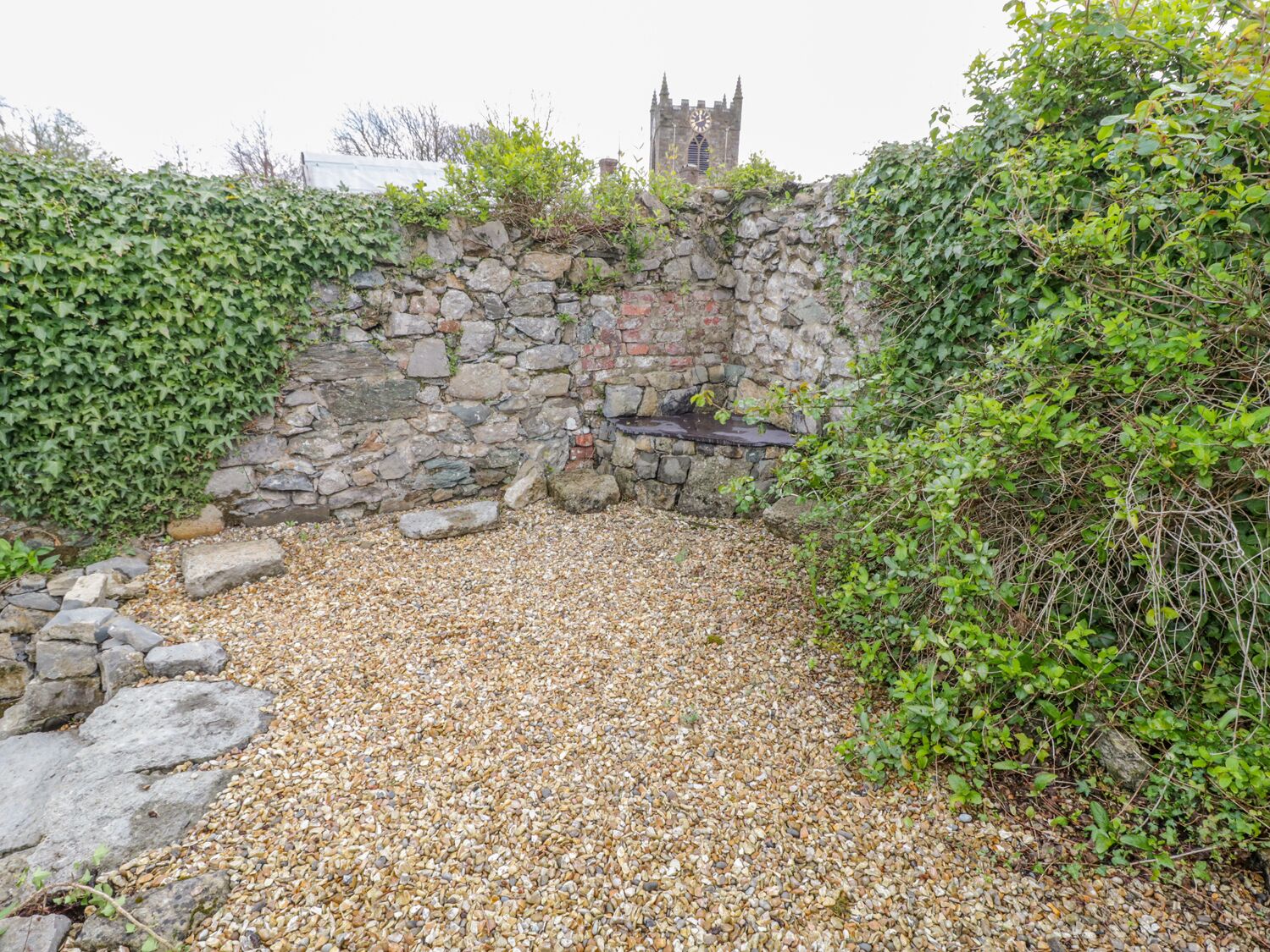 A stone wall with a clock tower in the background, surrounded by greenery.