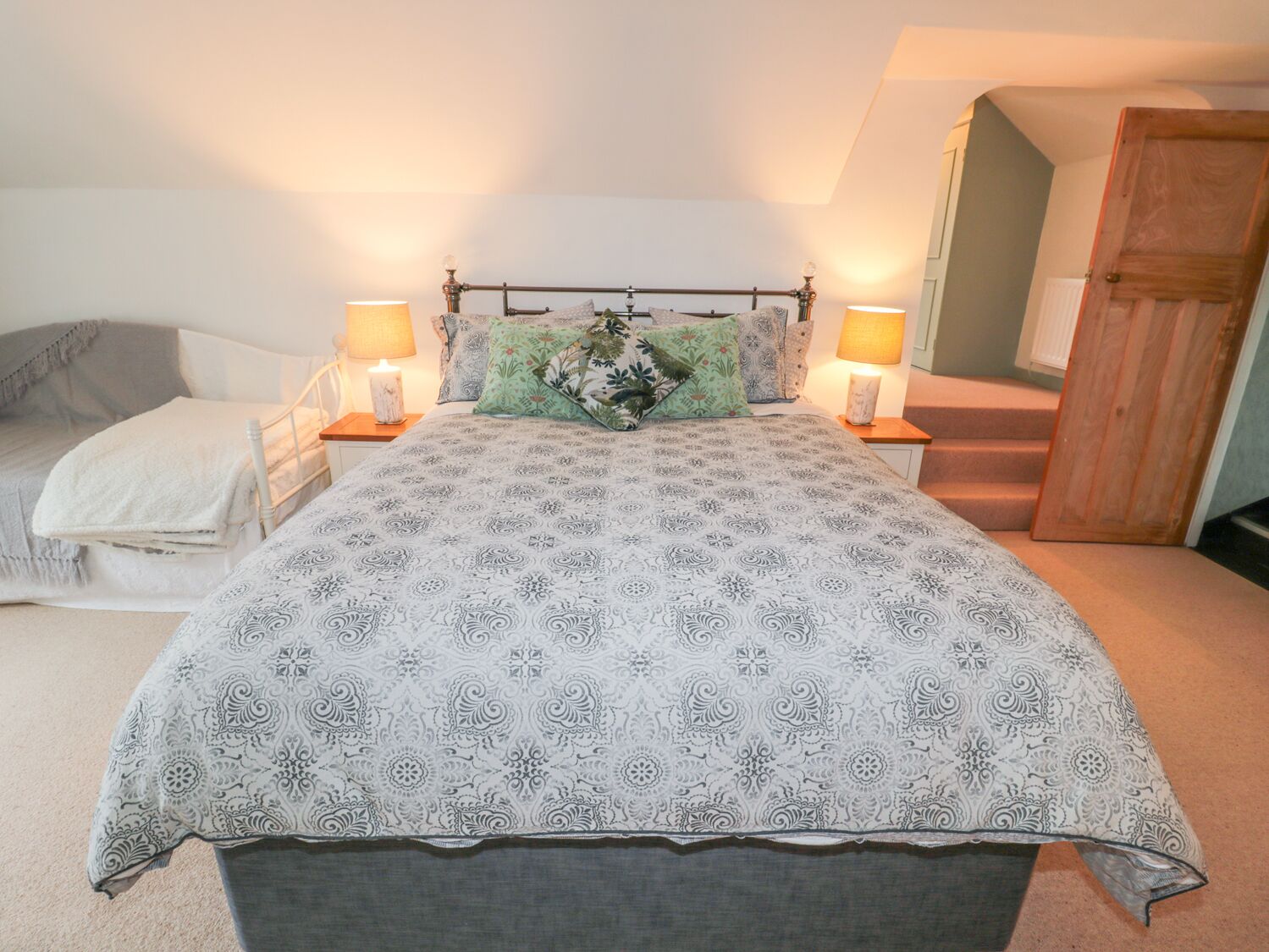 A bedroom with a bed, two bedside lamps, a nightstand, a chair, and a wooden door.