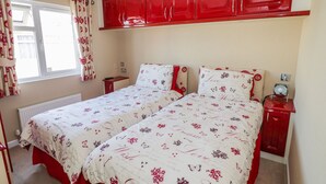 A bedroom with two beds, red cabinets above, a window with curtains, and a bedside table with a lamp. - Booster's Lodge (Stratford-upon-Avon)
