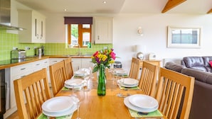 A dining area with a wooden table set for four, surrounded by wooden chairs. A vase with flowers is placed on the table. - Kingcup Cottage (St. Columb Major)