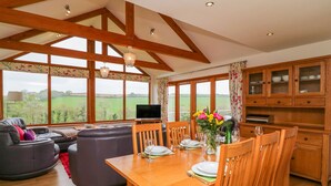 A spacious living area with a wooden dining table, comfortable seating, and large windows offering a view of the countryside. - Kingcup Cottage (St. Columb Major)