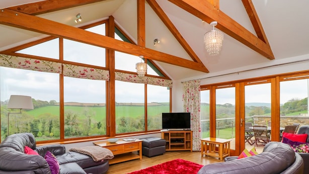 A spacious living room with a large TV, leather sofas, a red rug, and a balcony with a view of the countryside. - Kingcup Cottage (St. Columb Major)