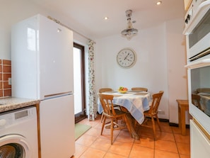 A compact kitchen with a washing machine, a dining table with chairs, a clock, and a ceiling fan. - Sarah's Cottage (Padstow)