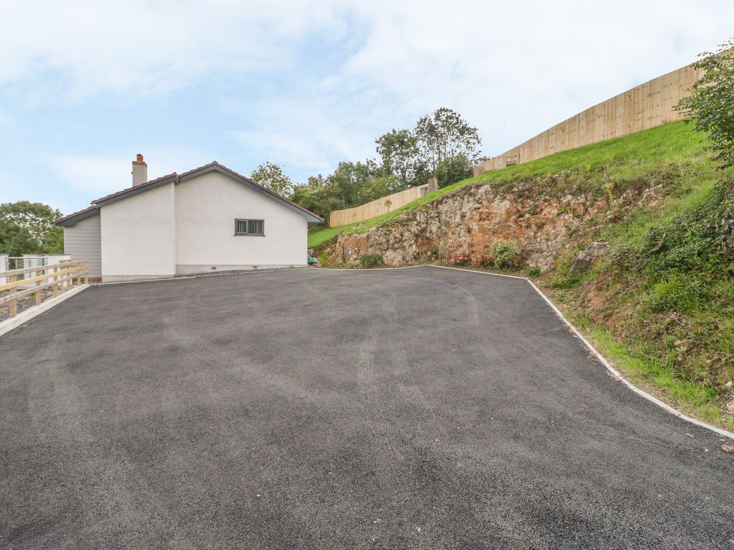 A paved driveway leading to a white building with a small window, surrounded by a grassy area and a stone wall.