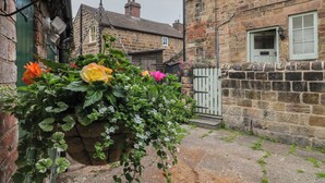 A stone building with a wooden door and a small garden with flowers. - Milford Cottage (Belper)